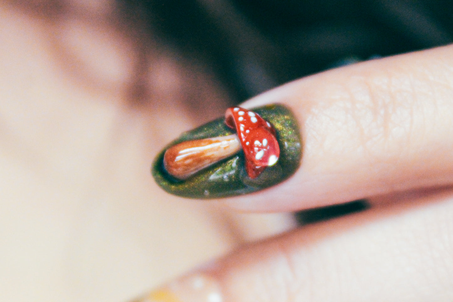 Close-up of the index finger wearing an Enchanted Pond press-on nail, featuring a sculpted red and beige mushroom on a deep green base with a subtle gold shimmer.