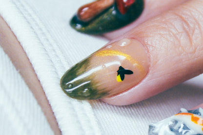 Close-up of the middle finger wearing an Enchanted Pond press-on nail, featuring raised dewdrops, a black firefly with a "glowing" yellow light, grassy green gradient, and a shimmering golden sunray detail.