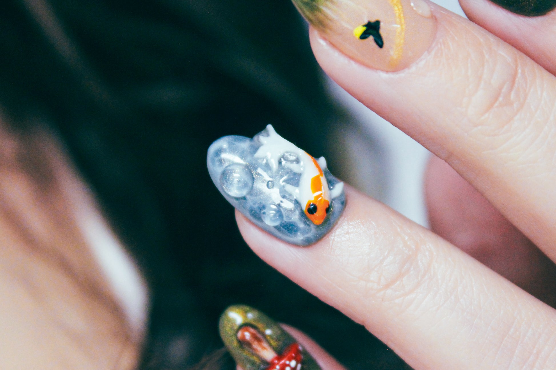 Close-up of the ring finger wearing an Enchanted Pond press-on nail, featuring a sculpted white and orange koi fish swimming through greenish-turquoise water ripples.