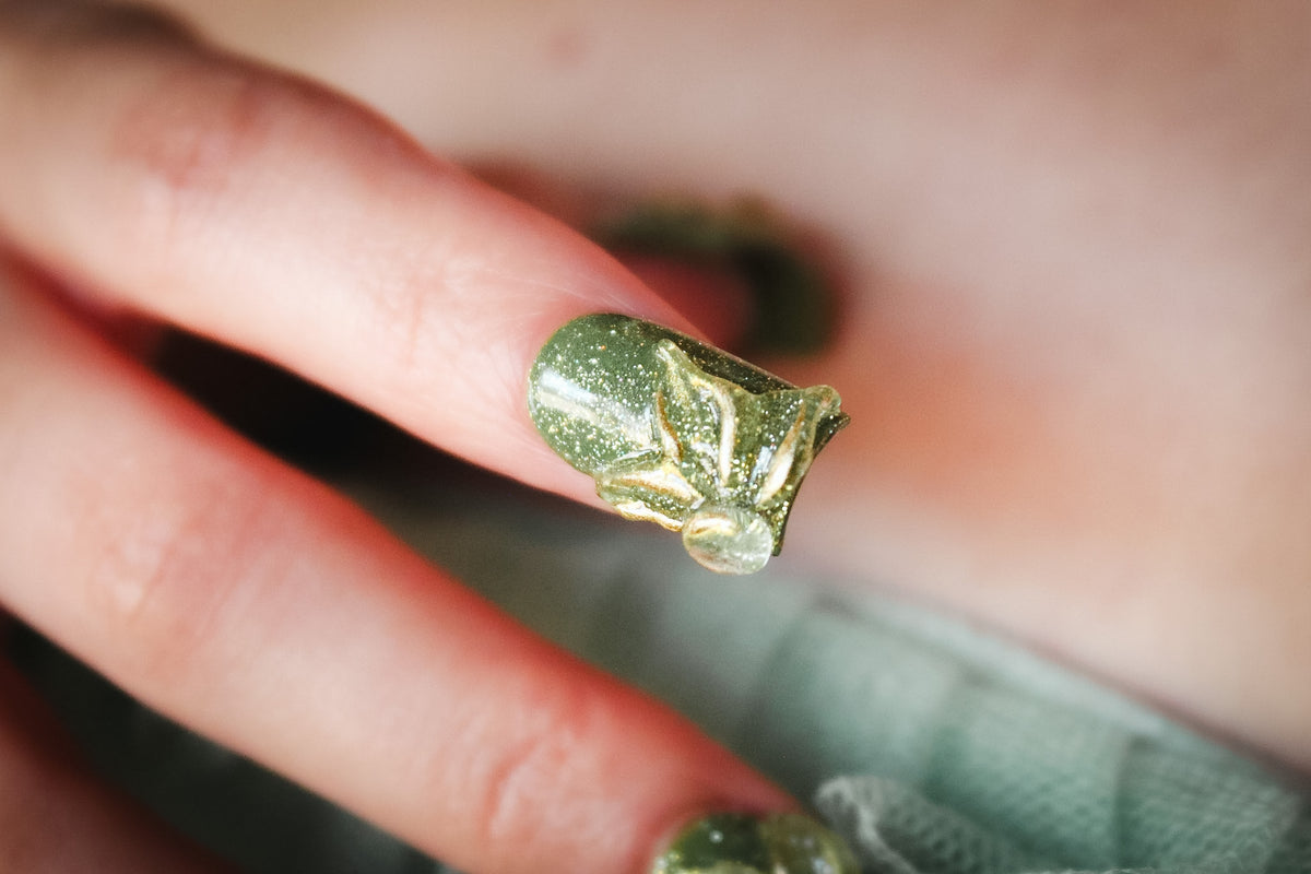 Close-up of a Green Blossom press-on nail with green glitter and a clear 3D flower outlined in gold.